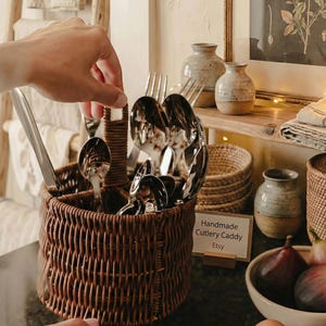 Puede incluir: Un cubertero tejido marrón con cucharas, tenedores y cuchillos. El cubertero tiene un asa y está dividido en secciones. Un letrero dice "Handmade Cutlery Caddy Etsy".