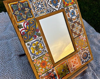 May include: A rectangular mirror with a wooden frame. The frame is decorated with colourful, patterned tiles. The tiles feature various designs in shades of blue, yellow, red, and green. The mirror is propped up on a wooden stand.