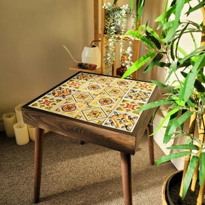 Op de afbeelding: Een vierkante bijzettafel met een donkerhouten frame en poten. Het tafelblad is bedekt met kleurrijke, patroon tegels met gele, oranje en blauwe ontwerpen. De tafel is ongeveer 61 cm hoog.