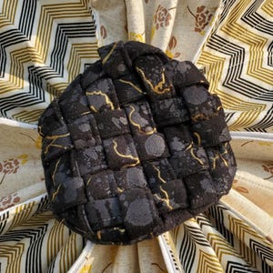 Puede incluir: Un cuenco de tela tejida con un patr&oacute;n moteado en negro y gris oscuro con finas l&iacute;neas met&aacute;licas doradas. El cuenco est&aacute; centrado sobre una base de tela plegada con fondo crema, detalles florales amarillos y un patr&oacute;n de chevr&oacute;n negro y oro.