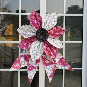 May include: A large, fabric flower decoration with alternating petals of patterned white and magenta fabric. The center is a dark, textured material. The flower is attached to a window with a white frame. The petals have a floral pattern.