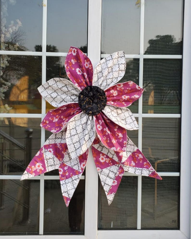 May include: A large, fabric flower decoration with alternating petals of patterned white and magenta fabric. The center is a dark, textured material. The flower is attached to a window with a white frame. The petals have a floral pattern.