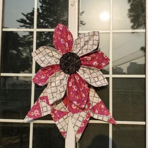 May include: A large, decorative fabric flower with layered petals. The petals alternate between a patterned cream and a floral print in shades of pink and red. The center is a dark, textured material. The flower is mounted on a white surface.