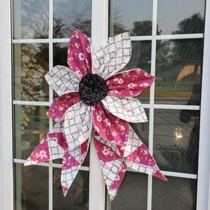 May include: A large, decorative fabric flower with alternating petals of patterned white and magenta fabric. The flower has a dark, textured center and is attached to a window. The flower is a unique home decor item.