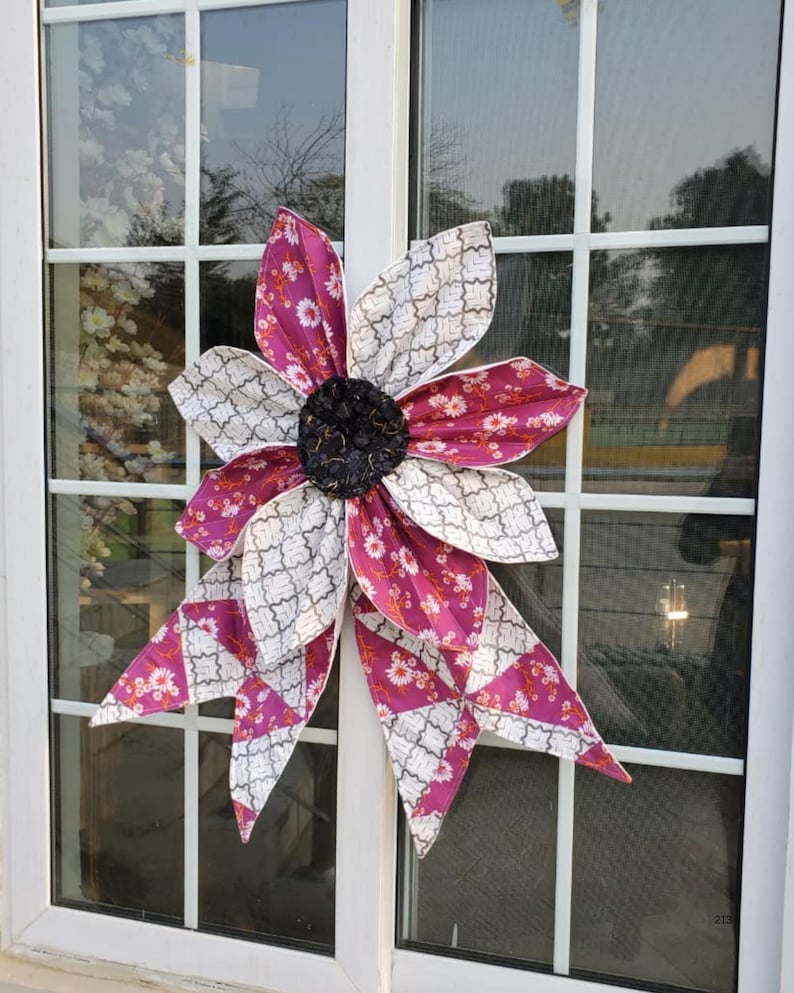 May include: A large, decorative fabric flower with alternating petals of patterned white and magenta fabric. The flower has a dark, textured center and is attached to a window. The flower is a unique home decor item.
