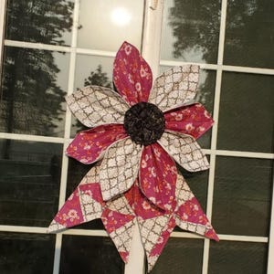 May include: A large, decorative fabric flower with alternating petals of patterned pink and cream fabric. The center is a dark, textured material. The flower is attached to a window, suggesting it's a home decor item.