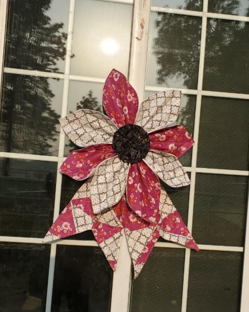 May include: A large, decorative fabric flower with alternating petals of patterned pink and cream fabric. The center is a dark, textured material. The flower is attached to a window, suggesting it's a home decor item.