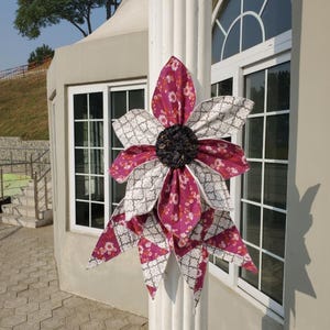 May include: A large, decorative fabric flower with layered petals in shades of burgundy, white, and black. The flower is attached to a white column, with a building and windows in the background. The petals have a floral and geometric pattern.