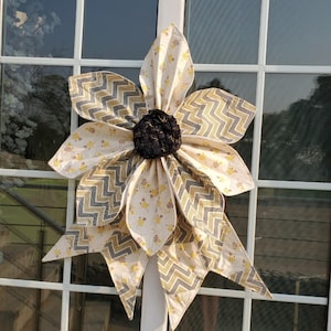 May include: A large, fabric sunflower decoration with a black center and layered petals. The petals feature a cream-colored fabric with yellow floral patterns and gray and gold chevron accents. The sunflower is mounted on a white window frame.