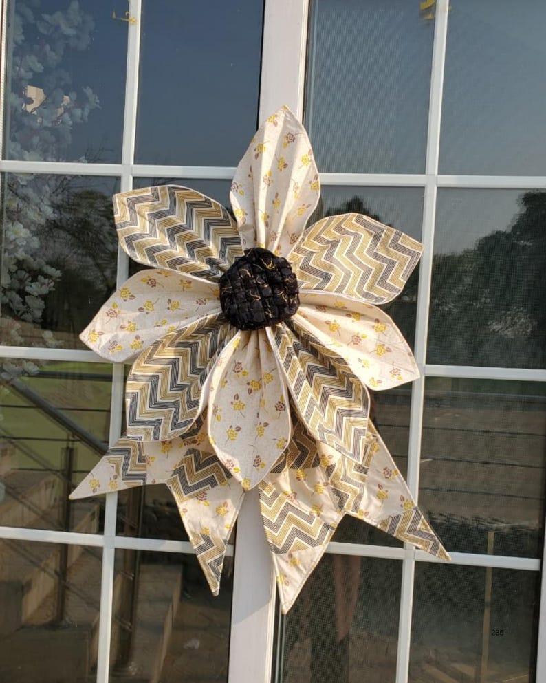 May include: A large, fabric sunflower decoration with a black center and layered petals. The petals feature a cream-colored fabric with yellow floral patterns and gray and gold chevron accents. The sunflower is mounted on a white window frame.
