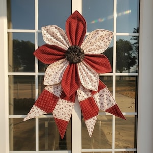 May include: A large, handmade fabric flower decoration with a red and white floral pattern. The flower has a dark brown center and a ribbon-like tail with a checkered design. It is displayed on a white window.