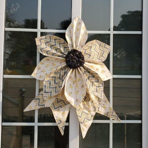 May include: A large, fabric flower decoration with a dark brown center and cream-colored petals. The petals feature a yellow and gray chevron pattern and small yellow floral accents. The flower is mounted on a white window frame.