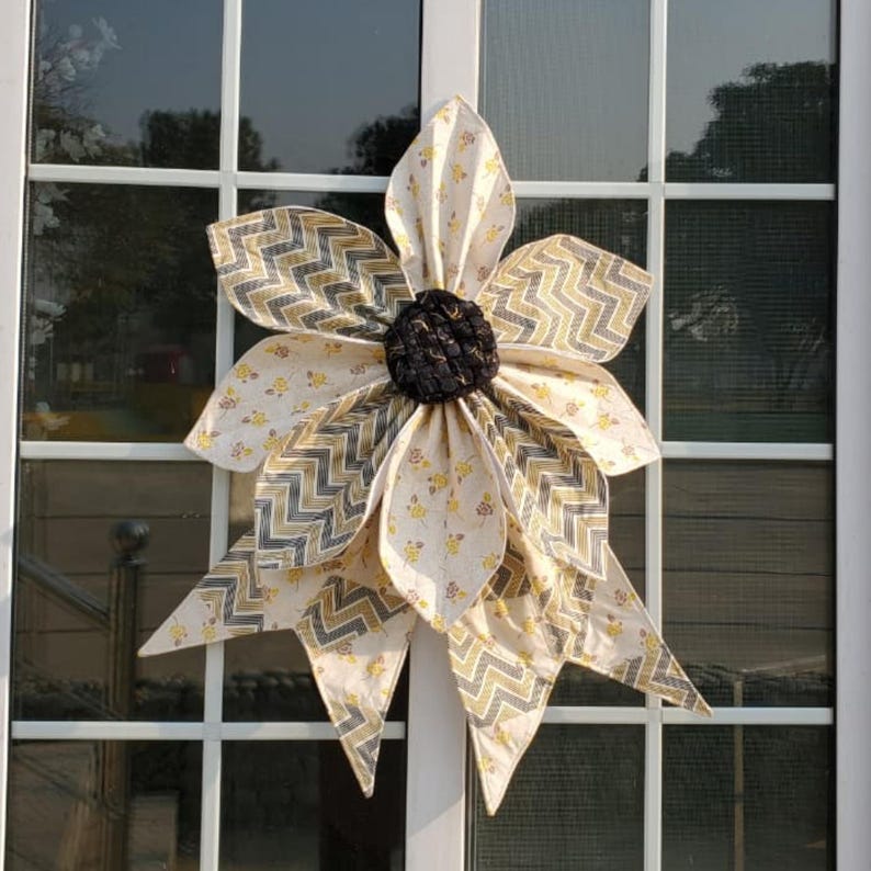 May include: A large, fabric flower decoration with a dark brown center and cream-colored petals. The petals feature a yellow and gray chevron pattern and small yellow floral accents. The flower is mounted on a white window frame.