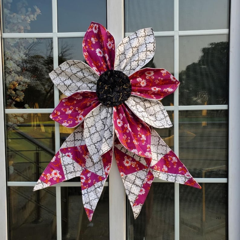 May include: A large, decorative flower made of fabric, featuring alternating petals of patterned white and magenta. The center is a textured black, and the flower is attached to a window. The petals have a floral pattern and a geometric design.