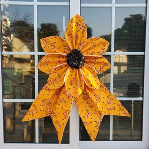 May include: A large, decorative fabric flower in shades of yellow and orange, with a dark center. The flower has multiple petals with a floral pattern and white trim. It is mounted on a window.