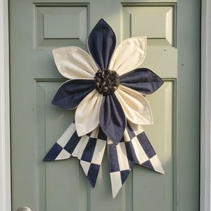 May include: A large, decorative flower-shaped wreath with a navy blue and cream color scheme. The flower petals alternate in color, with a dark center and a checkered bow. The wreath is hanging on a light green door.