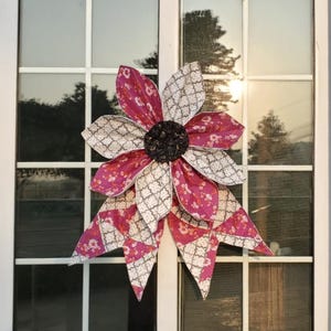 May include: A large, fabric flower decoration with alternating petals of patterned white and magenta fabric. The center is a dark, textured material. The flower is mounted on a white window frame, reflecting the outdoor scenery.