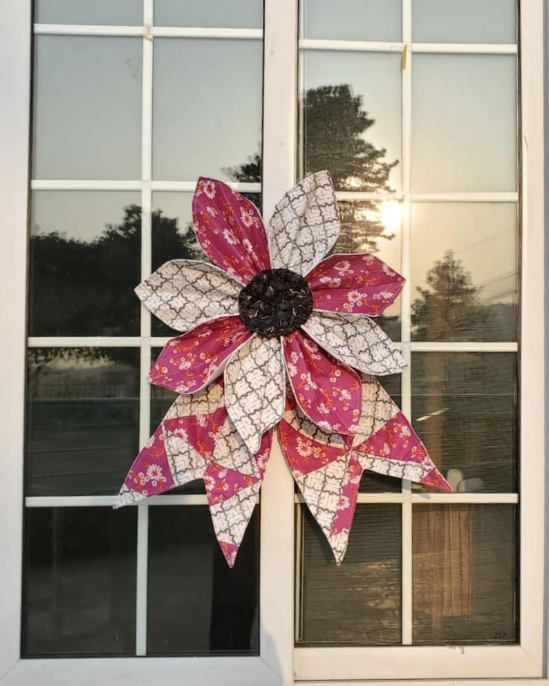 May include: A large, fabric flower decoration with alternating petals of patterned white and magenta fabric. The center is a dark, textured material. The flower is mounted on a white window frame, reflecting the outdoor scenery.