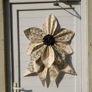 May include: A large, fabric flower decoration with layered petals in cream, yellow, and gray chevron patterns. The flower is centered with a dark, textured material and is mounted on a white door.