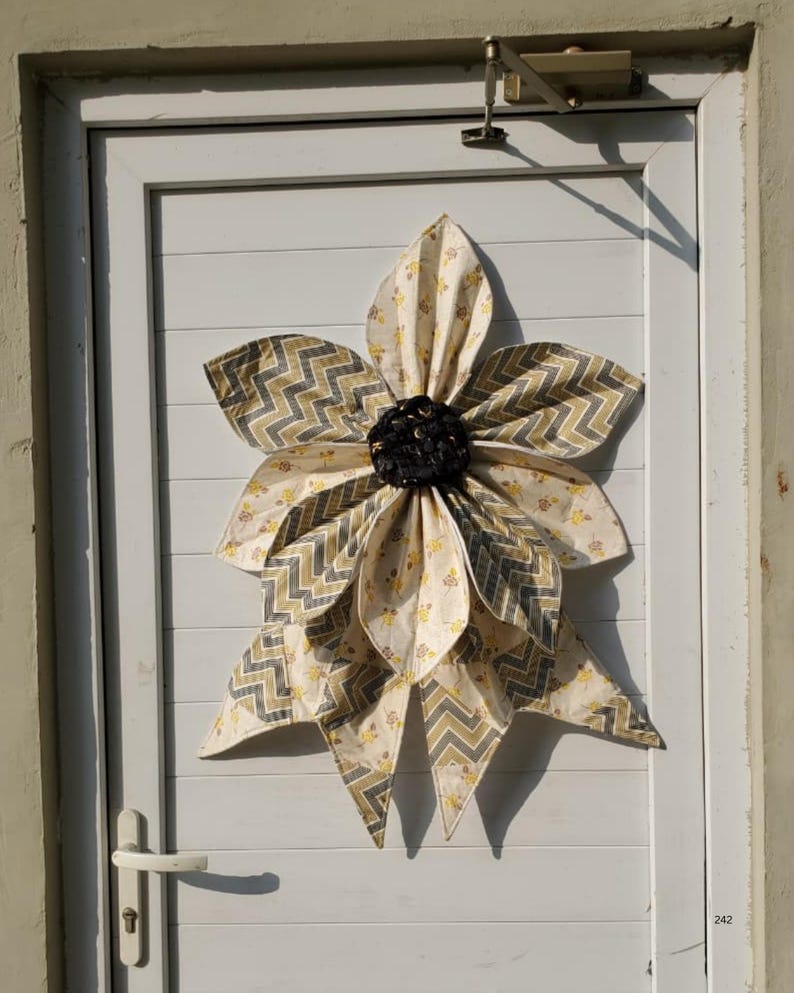 May include: A large, fabric flower decoration with layered petals in cream, yellow, and gray chevron patterns. The flower is centered with a dark, textured material and is mounted on a white door.