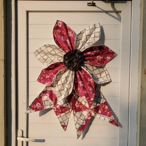 May include: A large, handmade fabric flower decoration with burgundy and white patterned petals. The center is a dark, textured material. The flower is mounted on a white door, creating a striking contrast.