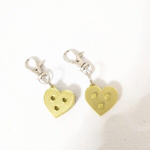 May include: Two gold-colored heart-shaped keychains with silver-colored clasps. Each heart has three small, evenly spaced holes. The keychains are displayed on a white surface.
