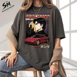 May include: A charcoal gray t-shirt featuring a graphic print of a red Honda NSX sports car and an anime-style character. The design includes the text FIFTH STAGE, HONDA NSX, and Japanese characters. The shirt has short sleeves and a crew neckline.