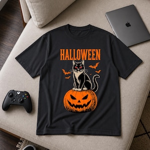 Puede incluir: Una camiseta negra de manga corta con un estampado gráfico de un gato negro con ojos rojos sentado sobre una calabaza naranja tallada. La palabra HALLOWEEN está impresa en naranja sobre el gato, rodeada de pequeños murciélagos voladores.