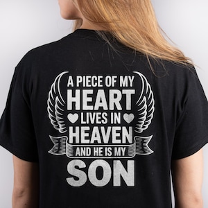 Puede incluir: Camiseta negra con un diseño gráfico blanco. El diseño incluye el texto "A PIECE OF MY HEART LIVES IN HEAVEN AND HE IS MY SON" con acentos de corazón y alas. La camiseta es de un material suave.