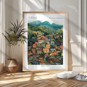 Op de afbeelding: Een ingelijste aquarelprint van Malang, Indonesië, met een dorp in de East Java Highlands. Het kunstwerk toont kleurrijke huizen met oranje daken, omringd door groene bomen en bergen. De print zit in een lichte houten lijst.