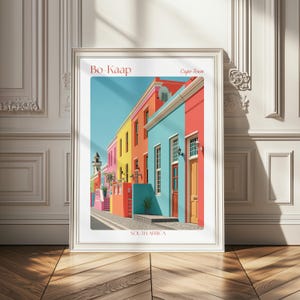 Può includere: Una stampa artistica incorniciata che raffigura una colorata scena di strada a Bo-Kaap, Città del Capo, Sudafrica. L'illustrazione mostra una fila di case vivaci nei toni del rosa, giallo, arancione e turchese sotto un cielo azzurro, con testo in alto e in basso.