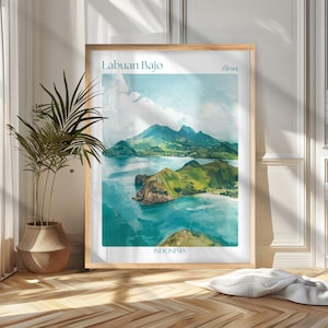 Op de afbeelding: Een ingelijste kunstdruk met een aquarel schilderij van Labuan Bajo, Indonesië. Het kunstwerk toont een kustlandschap met groene heuvels, turquoise water en de tekst "Labuan Bajo" en "Indonesia". De print zit in een houten lijst.