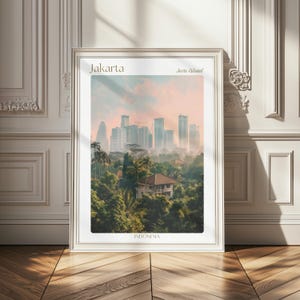 Op de afbeelding: Een ingelijste verticale wandkunstprint met het stadsgezicht van Jakarta op Java, Indonesië. De afbeelding toont een dicht groen bladerdak op de voorgrond met een moderne skyline van hoge gebouwen onder een zachtroze en oranje zonsondergang.