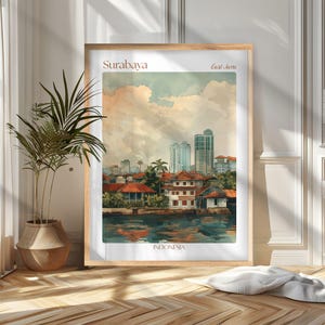 Op de afbeelding: Waterverfprint van Surabaya, Oost-Java, Indonesië, met een stadsgezicht met traditionele gebouwen en moderne wolkenkrabbers. Het kunstwerk is ingelijst in een licht houten frame en bevat de stadsnaam bovenaan.