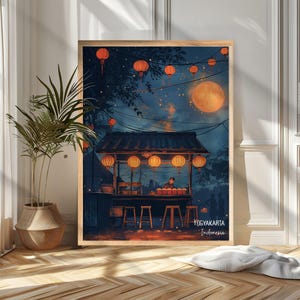 Könnte beinhalten: Ein gerahmter Kunstdruck zeigt eine Nachtszene eines Essensstandes in Yogyakarta, Indonesien. Das Kunstwerk zeigt orangefarbene Laternen, einen Vollmond und einen Verkäufer. Der Druck wird auf einem Holzboden neben einer Topfpflanze ausgestellt.