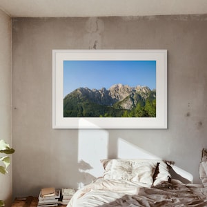 以下が含まれることがあります： 緑の木々と澄み切った青い空が広がる山脈の風景写真が額装されています。写真は寝室の灰色の壁に掛けられています。ベージュの掛け布団と枕が付いたベッドが前景にあります。