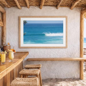 以下が含まれることがあります： 白い壁に飾られた、ターコイズブルーの海の波の額入りプリント。バーには、スツール、飲み物、パイナップルが置かれた木製のカウンターがあります。自然光が差し込み、リラックスした雰囲気を作り出しています。