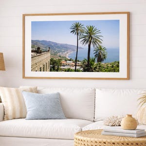 以下が含まれることがあります： ヤシの木、海岸沿いの町、青い空の下の海が描かれた額入りの風景画。白いソファとクッション、籐のコーヒーテーブルの上に飾られています。明るく風通しの良い部屋です。