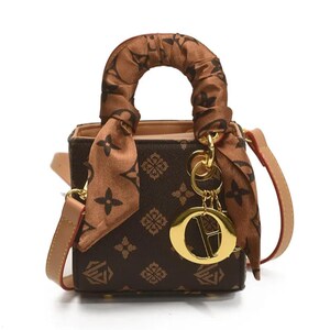 May include: A small brown handbag with a repeating pattern and a matching handle. The bag has a tan shoulder strap and a decorative scarf tied around the handle. A gold-colored charm hangs from the handle.