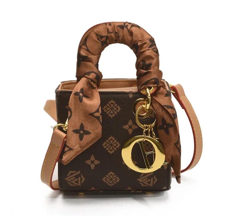 May include: A small brown handbag with a repeating pattern and a matching handle. The bag has a tan shoulder strap and a decorative scarf tied around the handle. A gold-colored charm hangs from the handle.