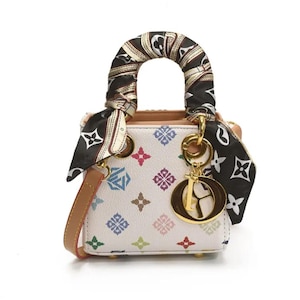 May include: A small, white handbag with a colorful geometric pattern. The bag features a brown handle wrapped with a patterned scarf and a gold-tone charm. A brown shoulder strap is also included.