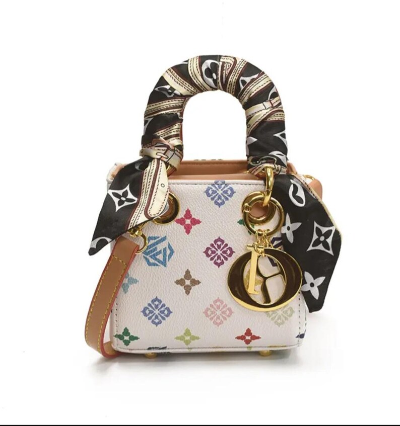 May include: A small, white handbag with a colorful geometric pattern. The bag features a brown handle wrapped with a patterned scarf and a gold-tone charm. A brown shoulder strap is also included.