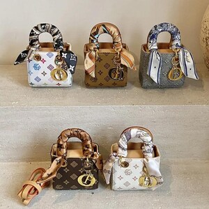 May include: A collection of miniature handbags in various colors, including white, brown, and gray. Each bag features a patterned design, a top handle, and a decorative scarf. Gold-colored charms and hardware add detail.