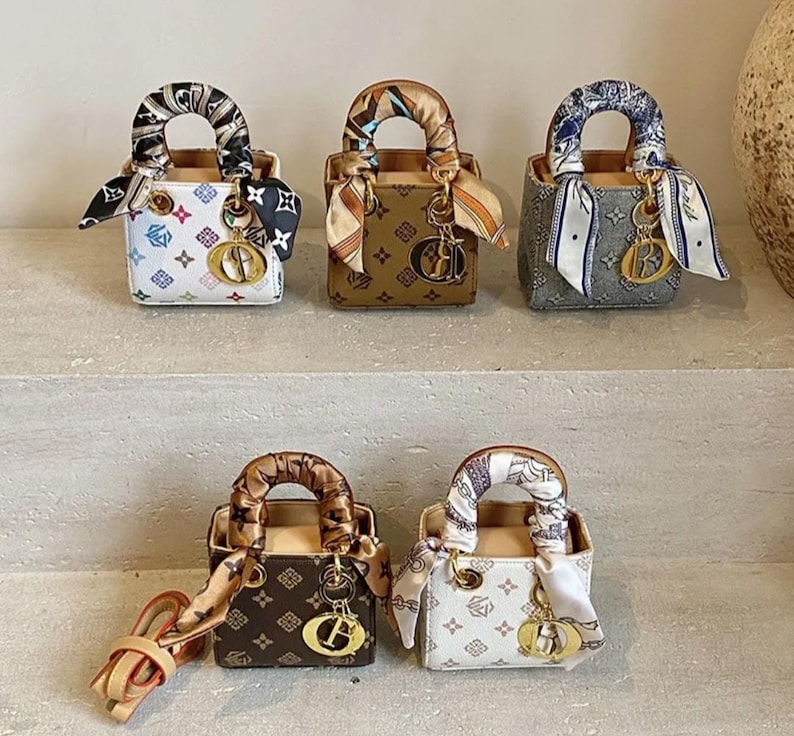 May include: A collection of miniature handbags in various colors, including white, brown, and gray. Each bag features a patterned design, a top handle, and a decorative scarf. Gold-colored charms and hardware add detail.