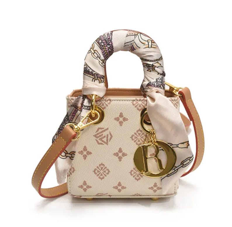 May include: A small, cream-colored handbag with a decorative pattern and a scarf wrapped around the handle. The bag has a gold-colored chain strap and a charm with the letter "R". The bag is approximately 6 inches wide.