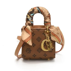 May include: Brown handbag with a repeating pattern and a decorative scarf wrapped around the handle. The bag features a gold-colored charm and a detachable shoulder strap. The bag is approximately 6 inches wide.