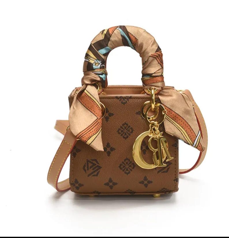 May include: Brown handbag with a repeating pattern and a decorative scarf wrapped around the handle. The bag features a gold-colored charm and a detachable shoulder strap. The bag is approximately 6 inches wide.