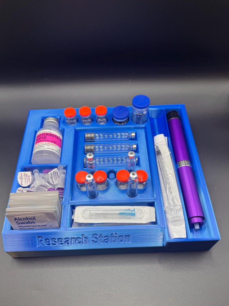 Puede incluir: Un organizador de estaci&oacute;n de investigaci&oacute;n de pl&aacute;stico azul con varios viales, jeringas y toallitas con alcohol. El organizador tiene compartimentos para diferentes art&iacute;culos, incluyendo un bol&iacute;grafo morado y botellas con tapas naranjas y azules. El texto "Research Station" es visible.