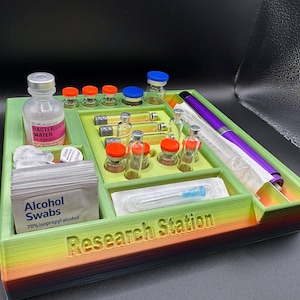 Puede incluir: Un organizador de estaci&oacute;n de investigaci&oacute;n multicompartimental en verde, amarillo y naranja. Contiene viales, botellas, toallitas con alcohol y un bol&iacute;grafo morado. El texto "Research Station" es visible.