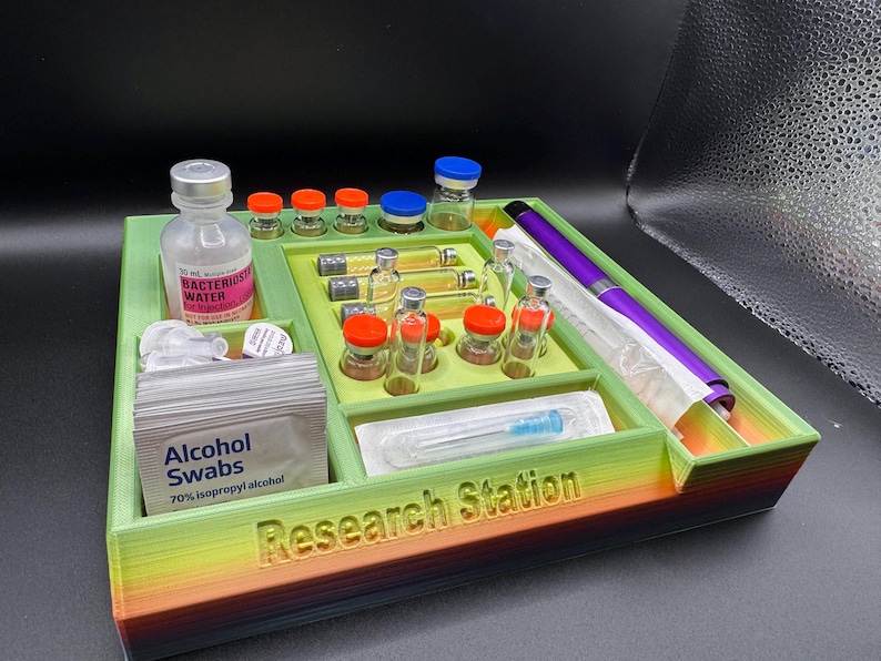 Puede incluir: Un organizador de estaci&oacute;n de investigaci&oacute;n multicompartimental en verde, amarillo y naranja. Contiene viales, botellas, toallitas con alcohol y un bol&iacute;grafo morado. El texto "Research Station" es visible.